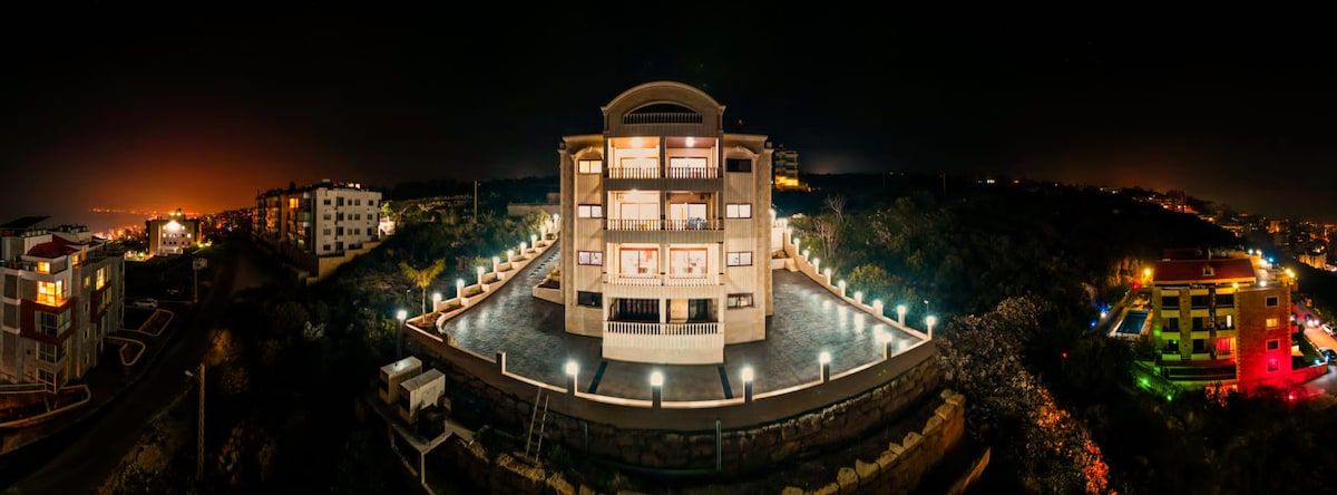 A modern building is presented in a panoramic night view, illuminated by exterior lights. A spacious outdoor area features pathways and landscaping, enhancing the architecture. Multiple balconies provide opportunities for enjoying the surrounding scenery, while neighboring structures contribute to the tranquil atmosphere.