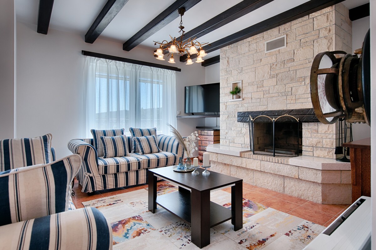 A comfortable living area is highlighted by a stone fireplace and wooden beams overhead. Light filters through sheer curtains, illuminating striped seating options. A modern television is mounted on the wall, while a decorative table sits on a colorful area rug.