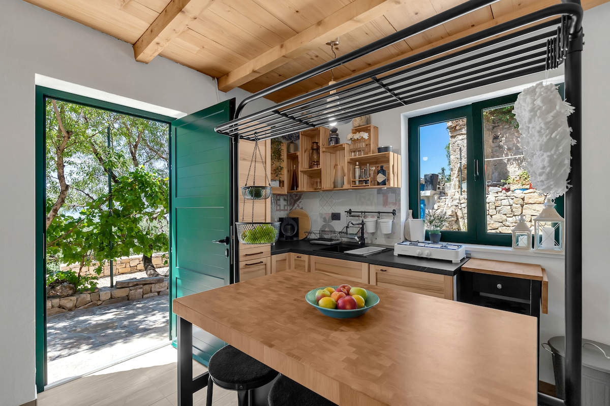 A well-equipped kitchen is visible, featuring a wooden dining table with a bowl of colorful fruit. Green doors frame the entrance to a serene outdoor view, while wooden cabinets and shelves offer storage and display space. Natural light enhances the warm, inviting atmosphere.