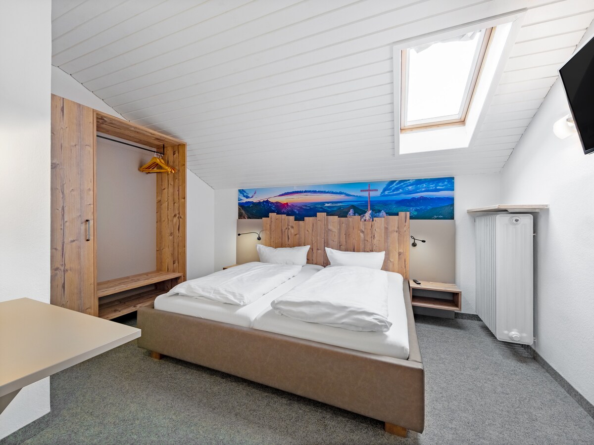 A well-appointed bedroom features a spacious bed with two separate bedding areas. Natural light filters in through a skylight above. Wooden accents are present on the headboard and wardrobe, while a mountain mural adds a scenic backdrop. A small table and television are visible.