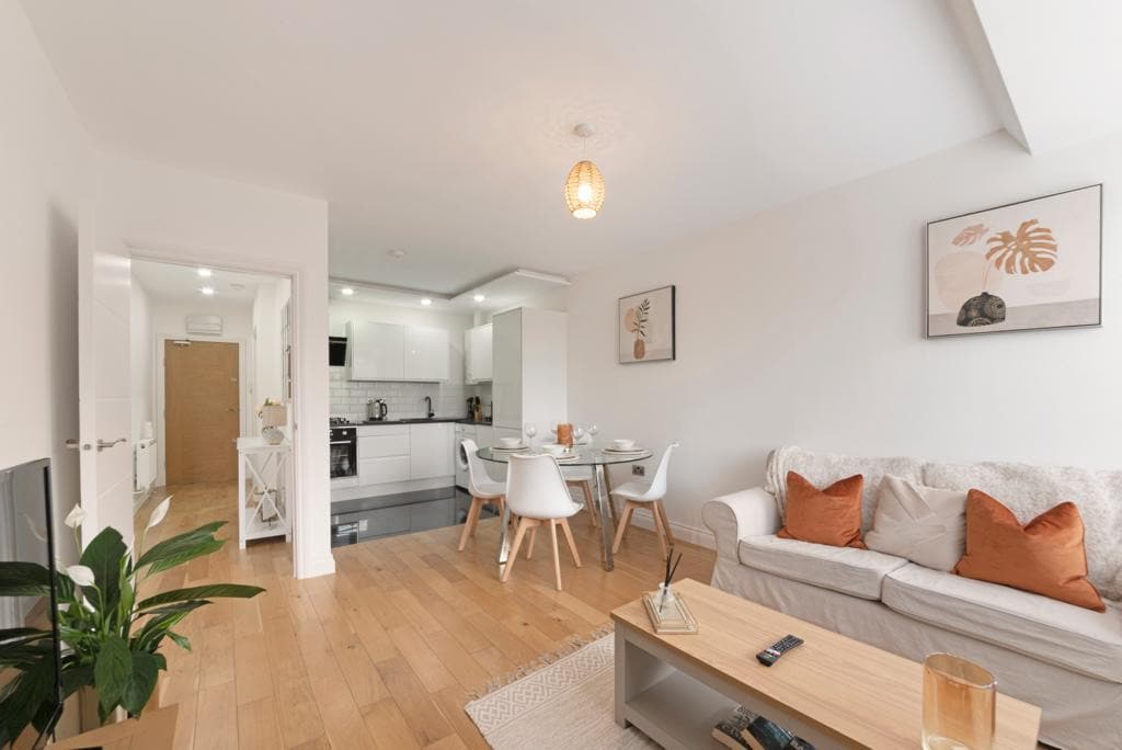 An open living space features light wood flooring and neutral-colored walls. A comfortable sofa with orange accent pillows is positioned next to a wooden coffee table. A dining area with a round table and four chairs is visible, as well as a modern kitchen in the background.