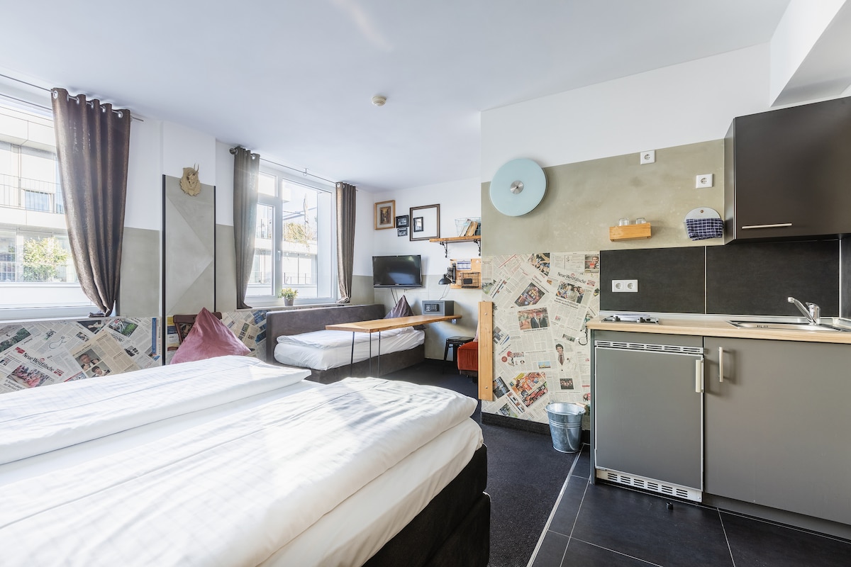 A studio apartment features a spacious bed and a kitchenette with modern appliances. Natural light fills the room through large windows adorned with neutral curtains. A seating area is visible, along with decorative wall art and a TV mounted on the wall.