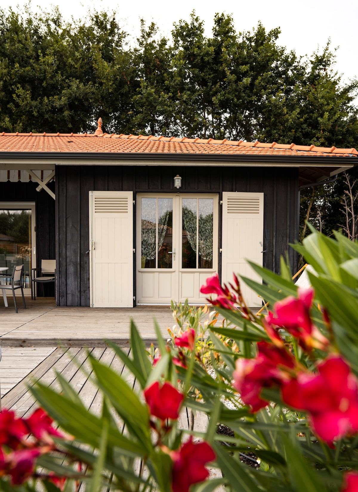 The exterior of the villa is framed by vibrant flowering plants. A set of double doors, adorned with clear glass panels, leads into the building. Light wooden shutters accentuate the windows, while a rustic tile roof adds charm to the wooden façade.