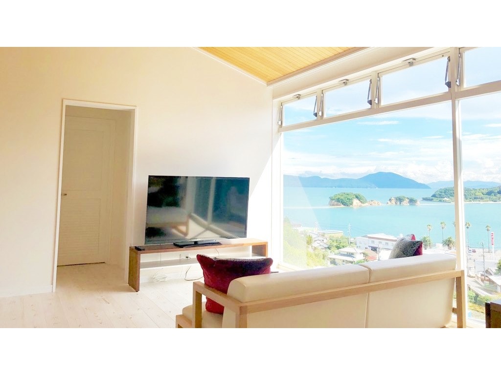 A bright living area features a large window overlooking the tranquil waters. A comfortable sofa is positioned in front of a television stand, creating a cozy space for relaxation. The warm wooden ceiling adds a touch of natural elegance to the room.