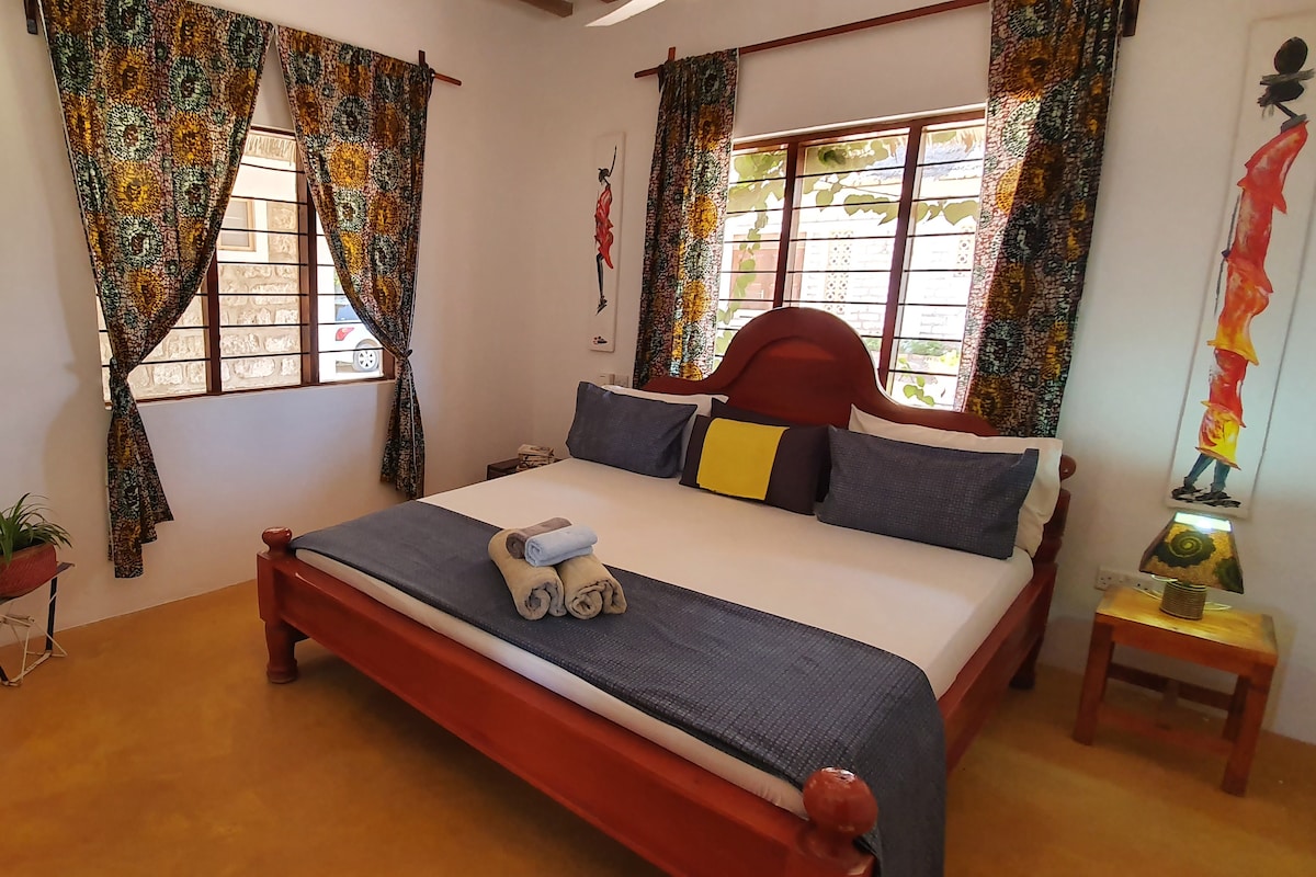 A spacious sleeping area features a wooden bed with a decorative headboard and a simple bedding arrangement. Colorful curtains frame the windows, allowing soft natural light to filter in. Decorative wall art and a peaceful ambiance enhance the inviting environment.
