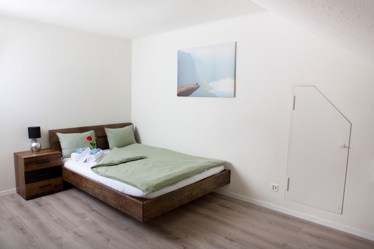 A spacious room is presented with a wooden bed featuring green bedding. A bedside table with a lamp and a decorative flower is included. A minimalist artwork adorns the wall, and a small door is visible, adding character to the space.