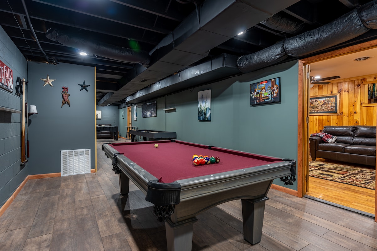 The game room features a billiards table with colorful balls arranged on a deep red felt surface. Dark walls provide a sleek backdrop, while low-hanging ceiling lights enhance the space. Nearby, framed artwork and wooden finishes add character to the inviting environment.