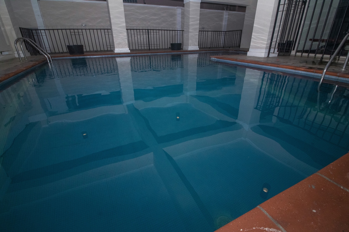 A communal swimming pool is presented, featuring a clear blue surface reflecting the surroundings. The pool is framed by a tiled edge, with safety rails visible on either side, providing a tranquil space for relaxation in the property.