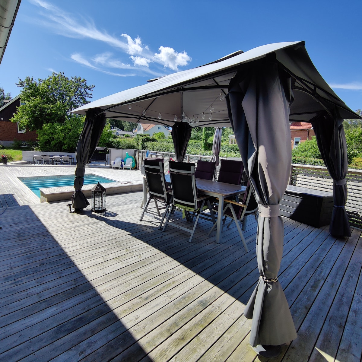 A shaded gazebo is shown on a wooden deck, providing a comfortable seating area with a table and several chairs. The pool area is visible in the background, surrounded by greenery under a bright blue sky.