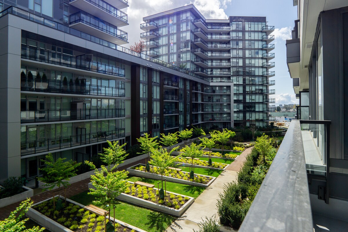 A modern residential complex is depicted, featuring several glass-fronted buildings surrounding a landscaped courtyard. The courtyard is adorned with neatly arranged greenery and trees, creating a serene outdoor space. A balcony railing is visible in the foreground, providing a view of the inviting landscape.