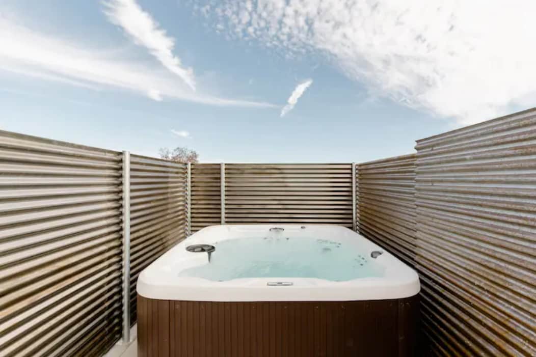 A hot tub is situated within a private fenced area, offering a tranquil space for relaxation. The tub features jets for soothing massage and is surrounded by weathered metal panels, complementing the open sky above. Clear blue skies and wispy clouds enhance the serene environment.