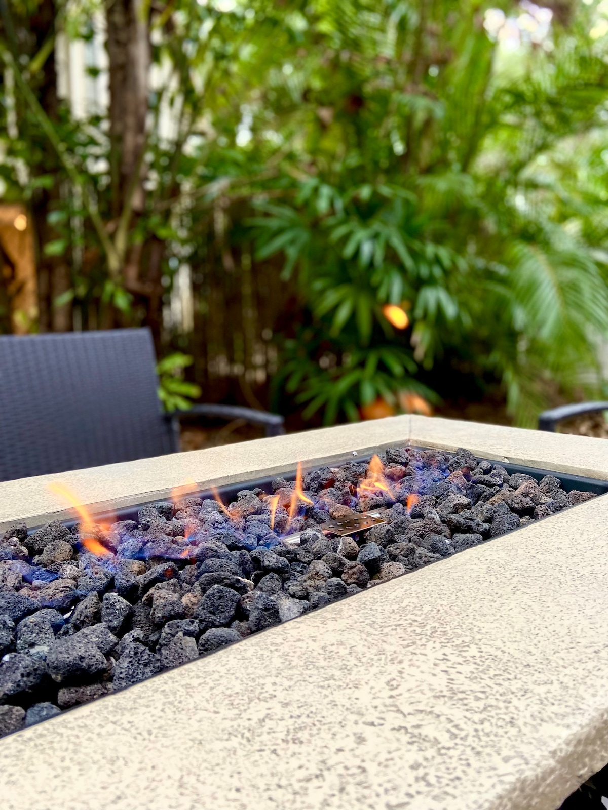 A fire pit surrounded by smooth stones emits gentle flames, providing a warm focal point in a lush backyard. Nearby, dark wicker chairs are positioned for relaxation among abundant greenery, creating a private and serene atmosphere.