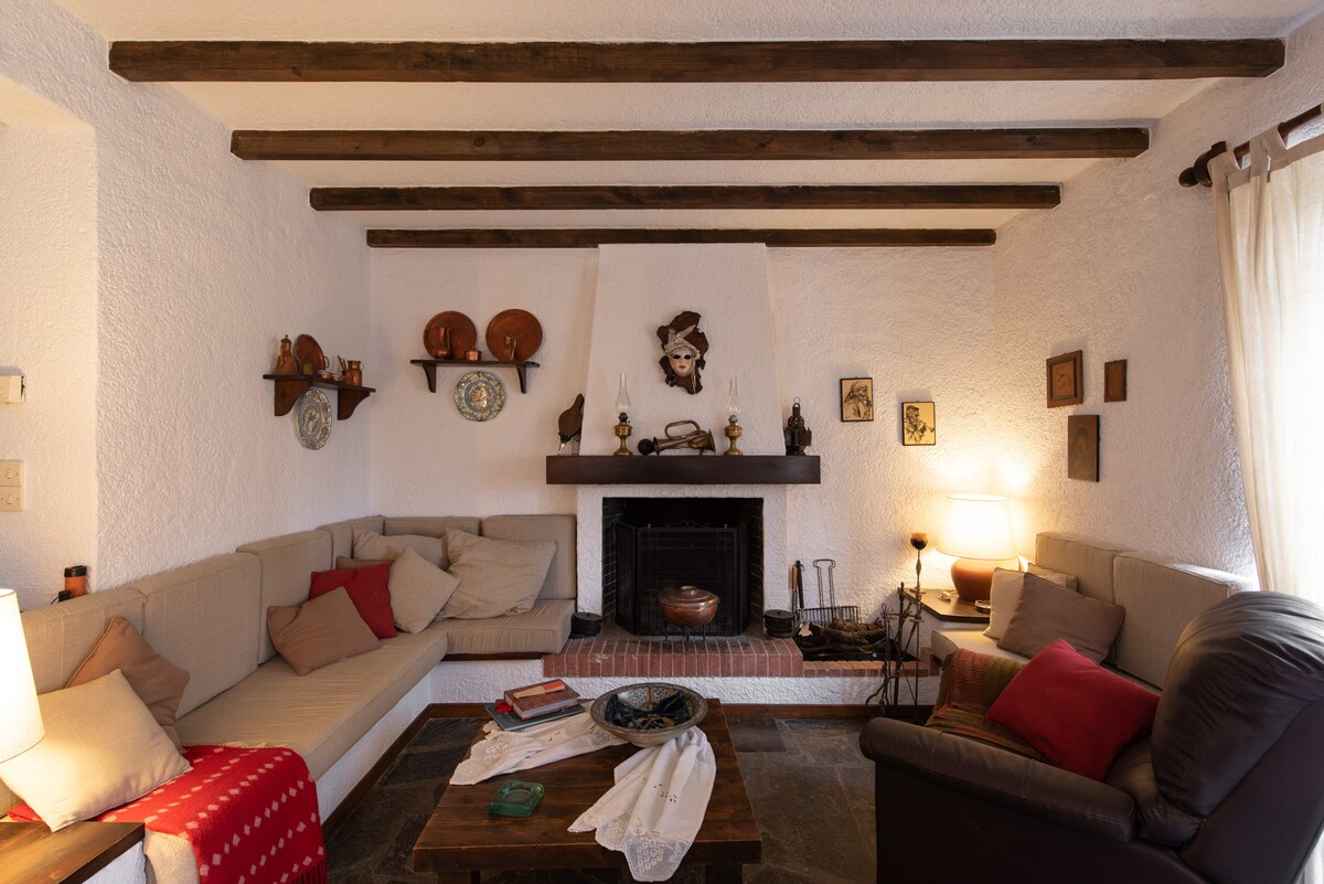 A cozy living area features a corner sofa with soft cushions, offering a relaxed seating option. A traditional fireplace is situated at the center, surrounded by decorative items on shelves. Light filters through the window, highlighting the wooden beams above and the warm tones of the decor.