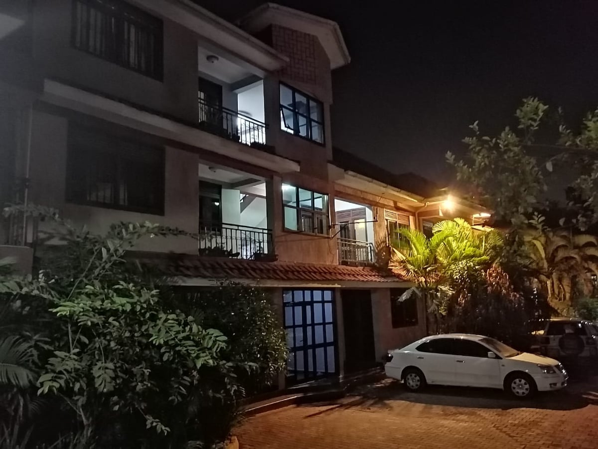 The exterior of a multi-story building is shown at night, featuring balconies and illuminated windows. Lush greenery surrounds the entrance, while a parked vehicle is visible in the foreground. Soft lighting highlights the architecture, creating a welcoming ambiance.