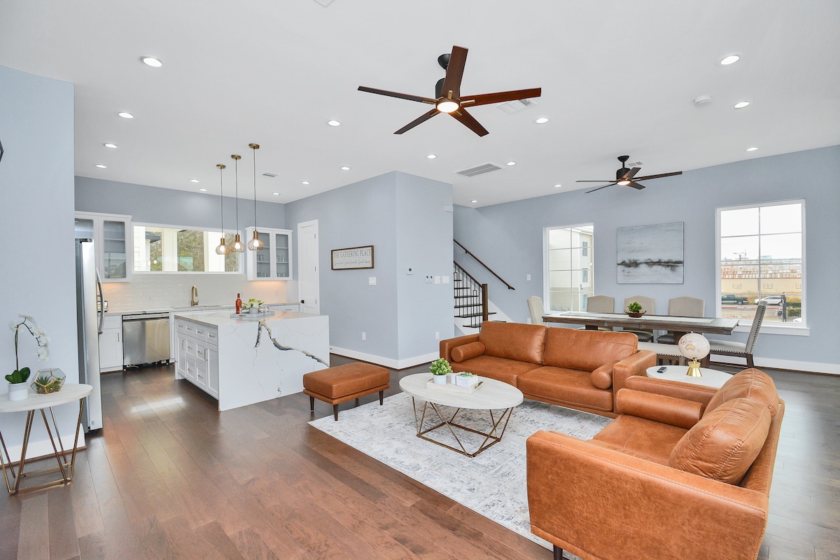 A spacious living area is shown with modern furniture arranged for comfort. A leather sofa and coffee table sit on a light area rug. The kitchen, with white cabinetry and stainless steel appliances, is visible in the background, complemented by contemporary lighting fixtures.