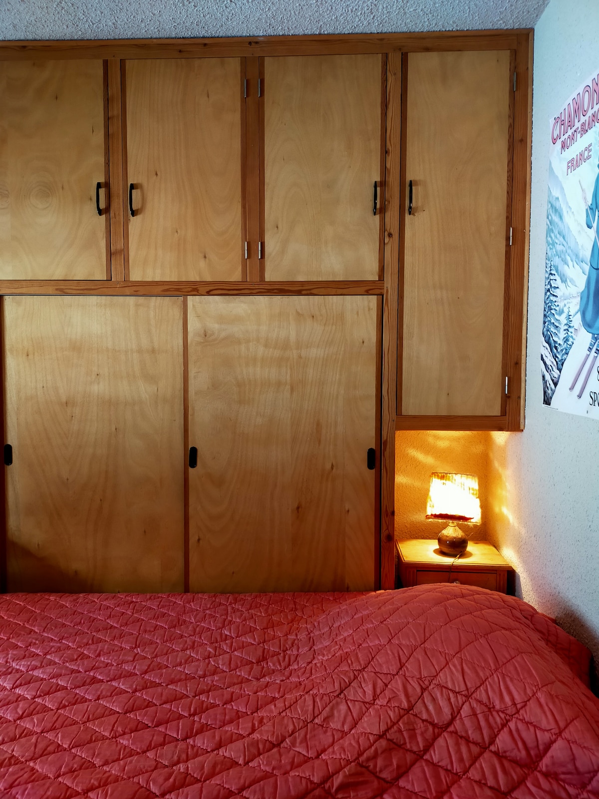 A cozy sleeping area is framed by warm wooden cabinetry above a spacious bed covered in a coral quilt. A small bedside lamp casts a soft glow, accentuating the simplicity of the space, while a vintage ski poster adds a subtle touch of charm.