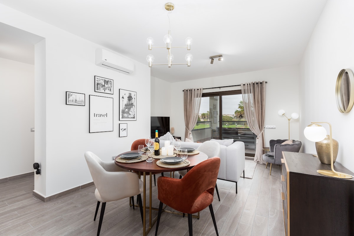 The dining area features a round table set for four, surrounded by stylish chairs in muted tones. A large window allows natural light to brighten the space, while contemporary artwork decorates the walls. The adjacent lounge area includes a comfortable sofa and a TV.