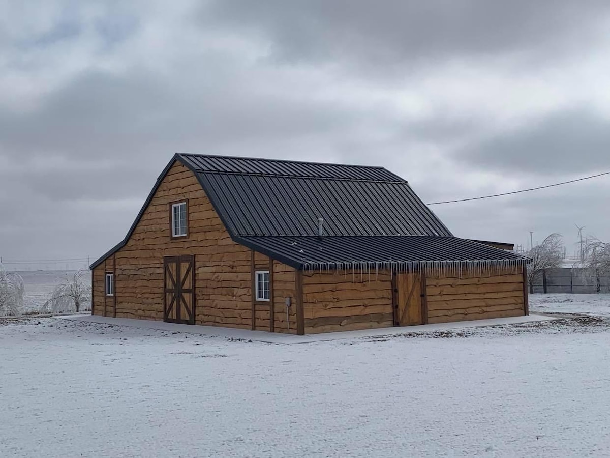 A newly remodeled barn-dominium is depicted, featuring a wooden exterior with a contrasting dark metal roof. Icicles hang from the eaves, while a light blanket of snow covers the ground. The structure displays a classic barn design with large doors and small windows.