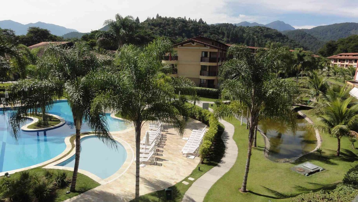 The image showcases a lush, green landscape featuring well-maintained gardens, palm trees, and a large swimming pool with a winding shape. Pathways are visible leading through the property, with residential buildings in the background and mountainous terrain providing a serene backdrop.