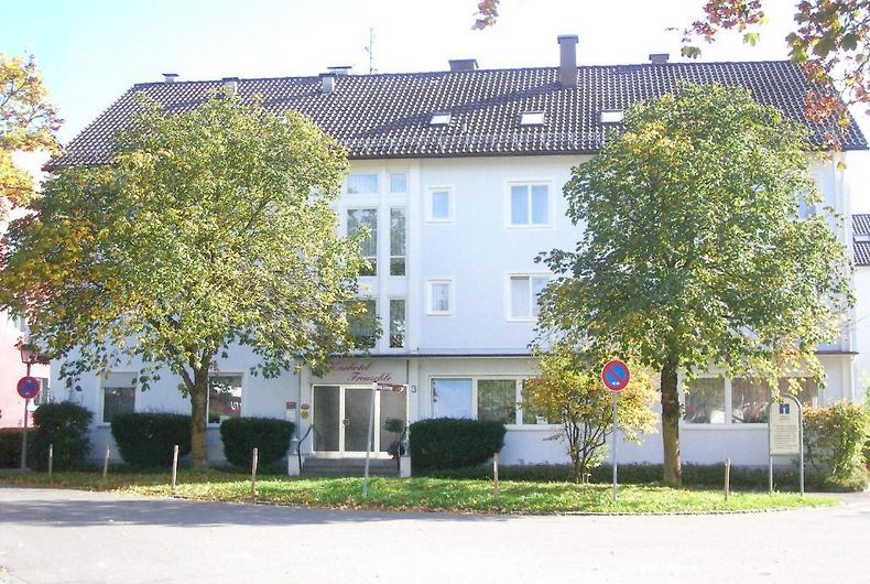 A four-story building is presented, featuring a light-colored facade with multiple symmetrical windows. Two large trees are positioned in front, providing greenery. The entrance is marked by a subtle sign, and a circular road with parking signs is visible in the foreground.
