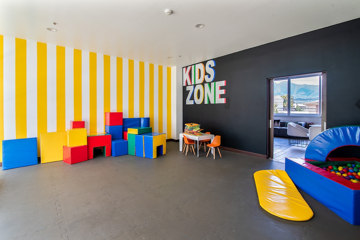 A colorful children's play area features striped yellow and white walls. Various soft foam blocks in primary colors are arranged throughout the space. A small table with chairs is positioned nearby, and a play structure with a ball pit is visible, creating a playful environment.