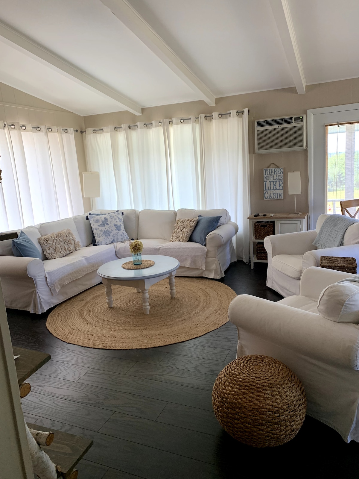 A light-filled living area features a comfortable white sectional sofa adorned with blue and patterned cushions. A circular coffee table is centered on a natural fiber rug. Sheer curtains frame large windows, allowing natural light to illuminate the space.