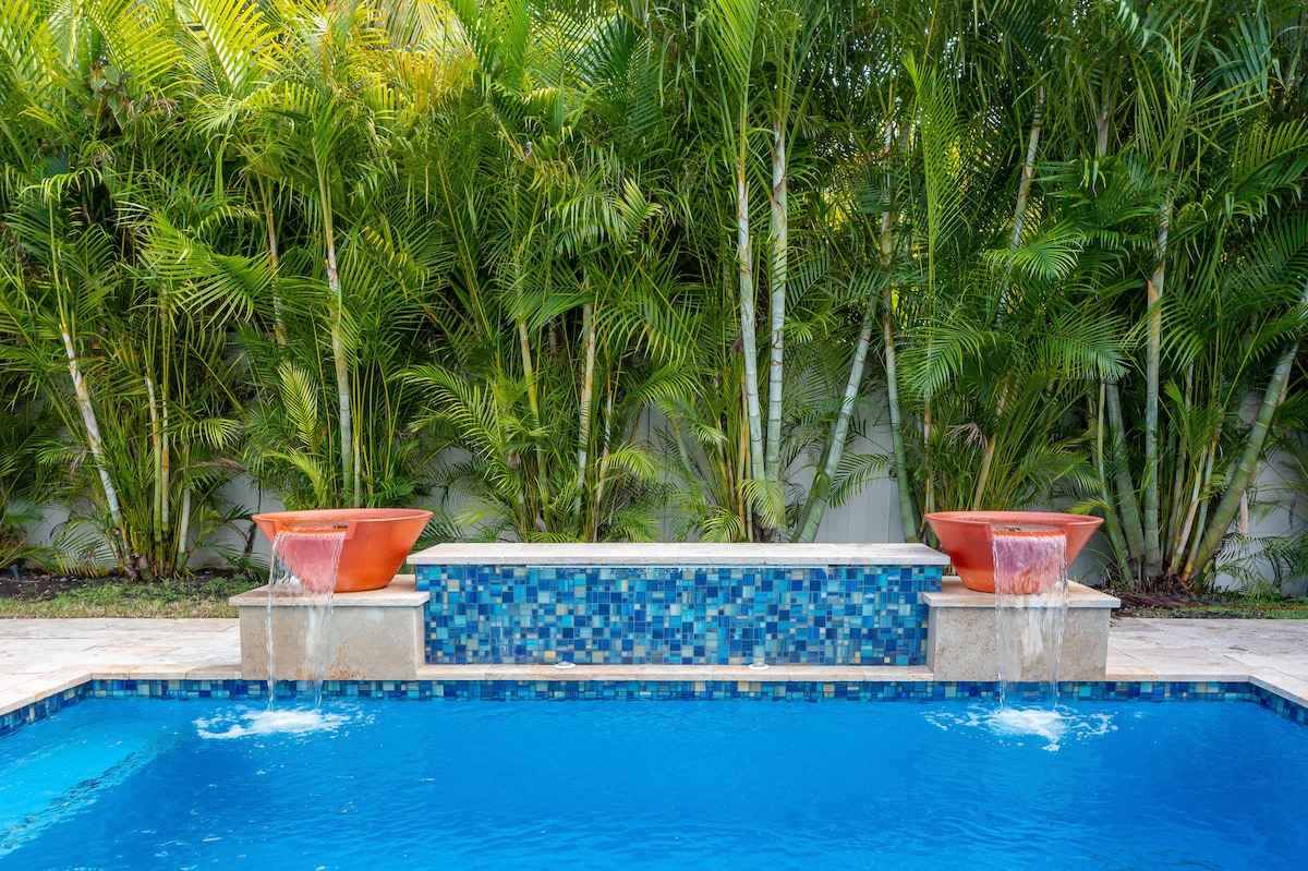 A heated salt-water pool is framed by lush tropical palms, providing a serene setting. Two decorative fountains are positioned at either end of a raised seating area, with vibrant blue tiles adding a touch of color to the inviting space.