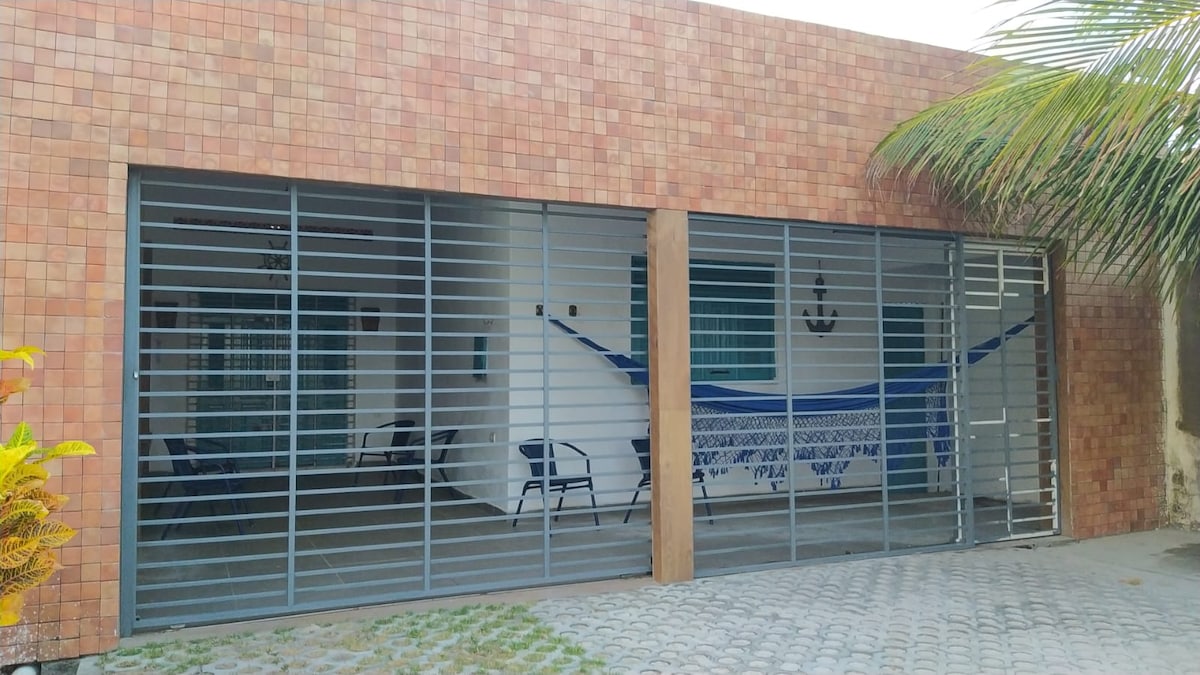 A spacious outdoor area is enclosed by metal slats, providing a sense of security. Two hammocks are positioned for relaxation, alongside several chairs. A textured brick wall complements the setting, while tropical greenery is seen in the foreground, enhancing the environment.