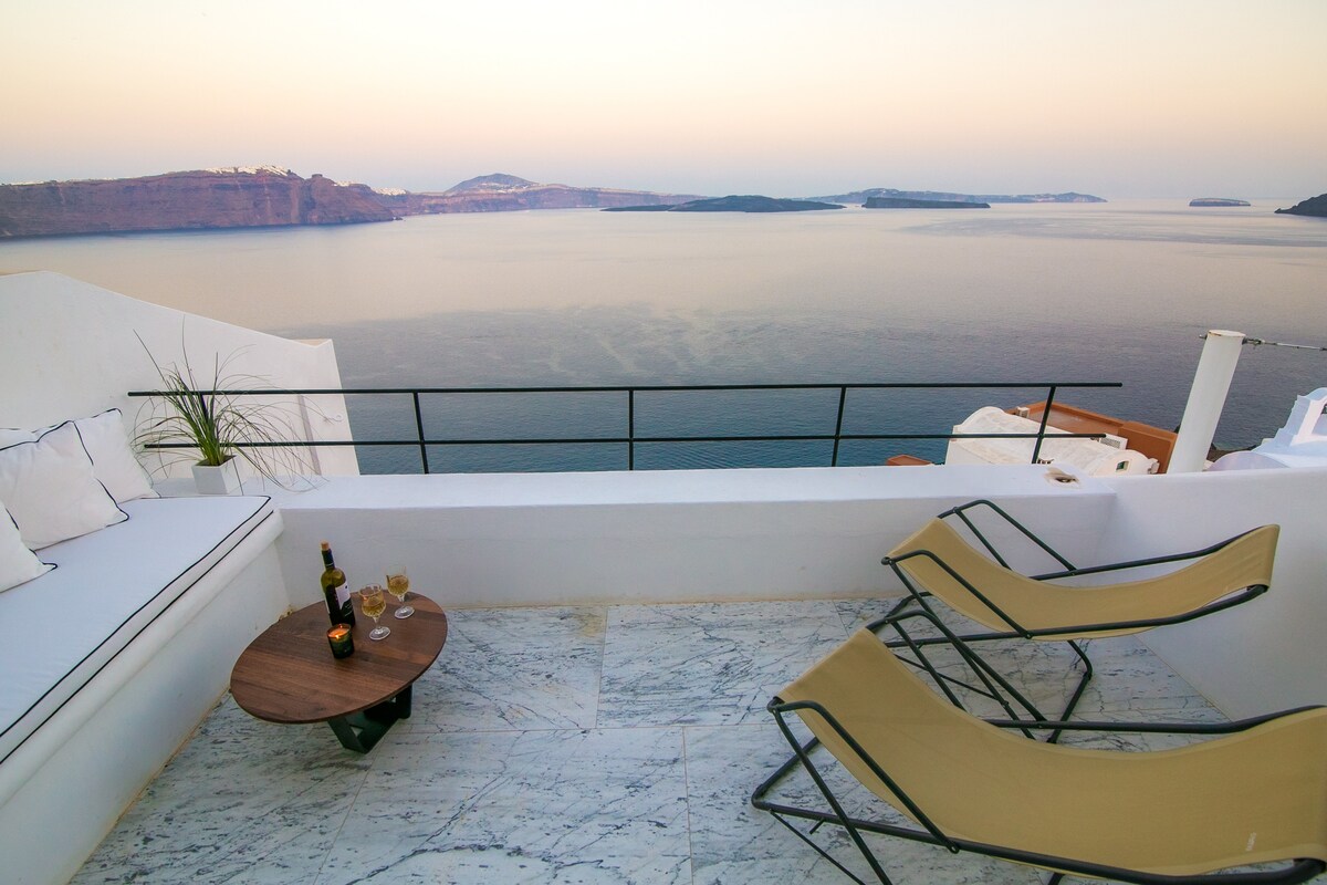 A private terrace is showcased with a marble surface and minimalistic furnishings. A set of sun loungers is positioned beside a low table, topped with drinks. The panoramic view includes calm waters and distant islands, illuminated by soft, fading light.