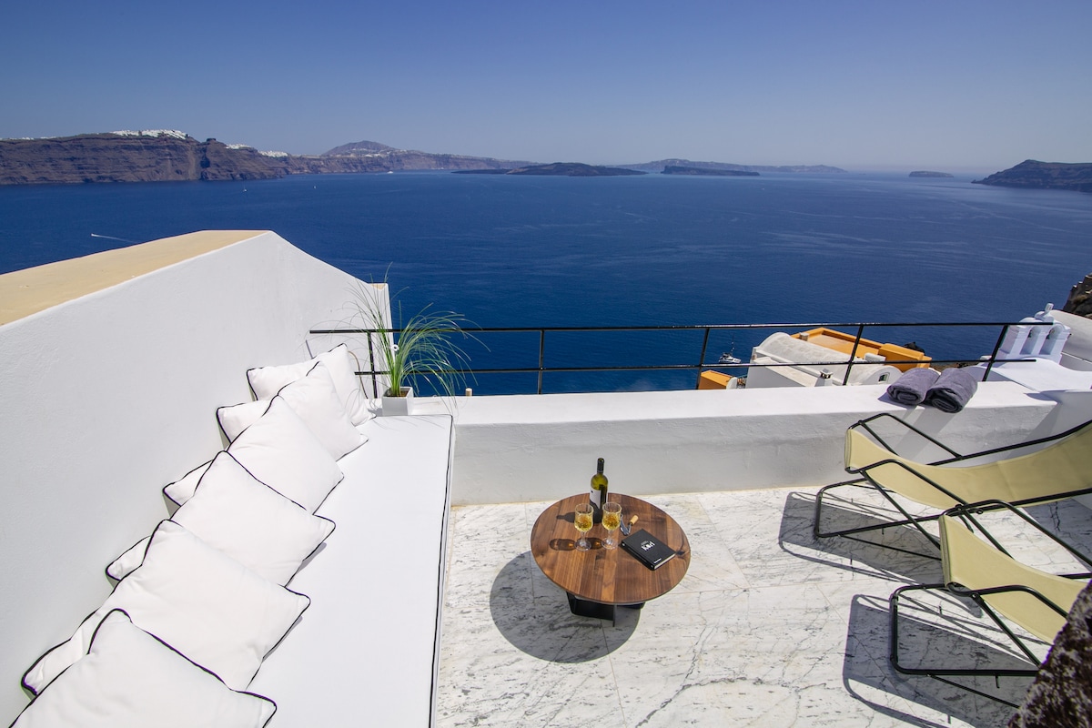 A panoramic terrace is presented with a round table and two lounge chairs overlooking the caldera. Cushioned seating enhances comfort, while a bottle of wine and two glasses are placed on the table, inviting relaxation. The expansive view captures the serene blue waters and distant islands.