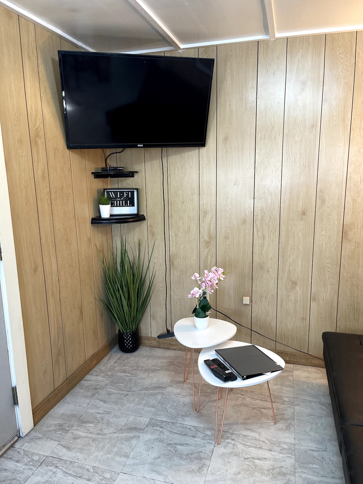A corner of a room is shown, featuring a mounted television above a small, white table. A laptop and remote control rest on the table, while a potted plant adds a touch of greenery. The walls are covered in wooden panels for a rustic feel.