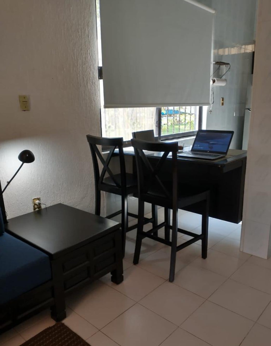 A compact workspace is featured, including a sleek desk with a laptop and two high-backed chairs. Natural light enters through a partially opened window, illuminating the minimalist decor and tiled floors.