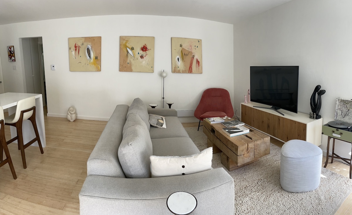 A modern living area features a spacious gray sectional sofa and a rustic wood coffee table. Abstract artwork adorns the wall, while a large flat-screen TV is positioned across from the seating. Various decorative elements, including a soft ottoman, enhance the inviting look.