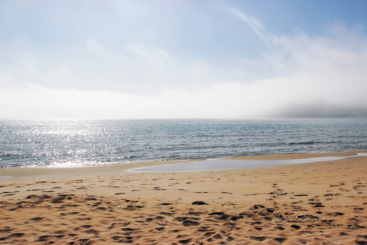 A sandy beach is seen stretching along the shore, meeting the tranquil sea. Gentle waves reflect sunlight, creating a shimmering surface. Mist is present in the distance over the water, contributing to a serene coastal atmosphere.