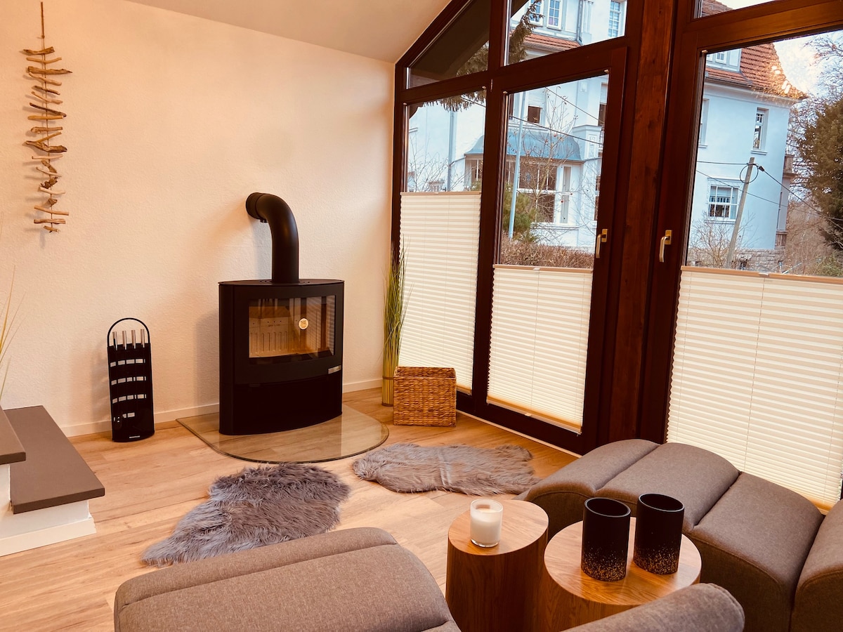 A cozy living area features large windows allowing natural light to enter. A modern fireplace is positioned against a light-colored wall, with two gray lounge chairs and a wooden side table nearby. Textured rugs and a candle add a touch of warmth to the space.