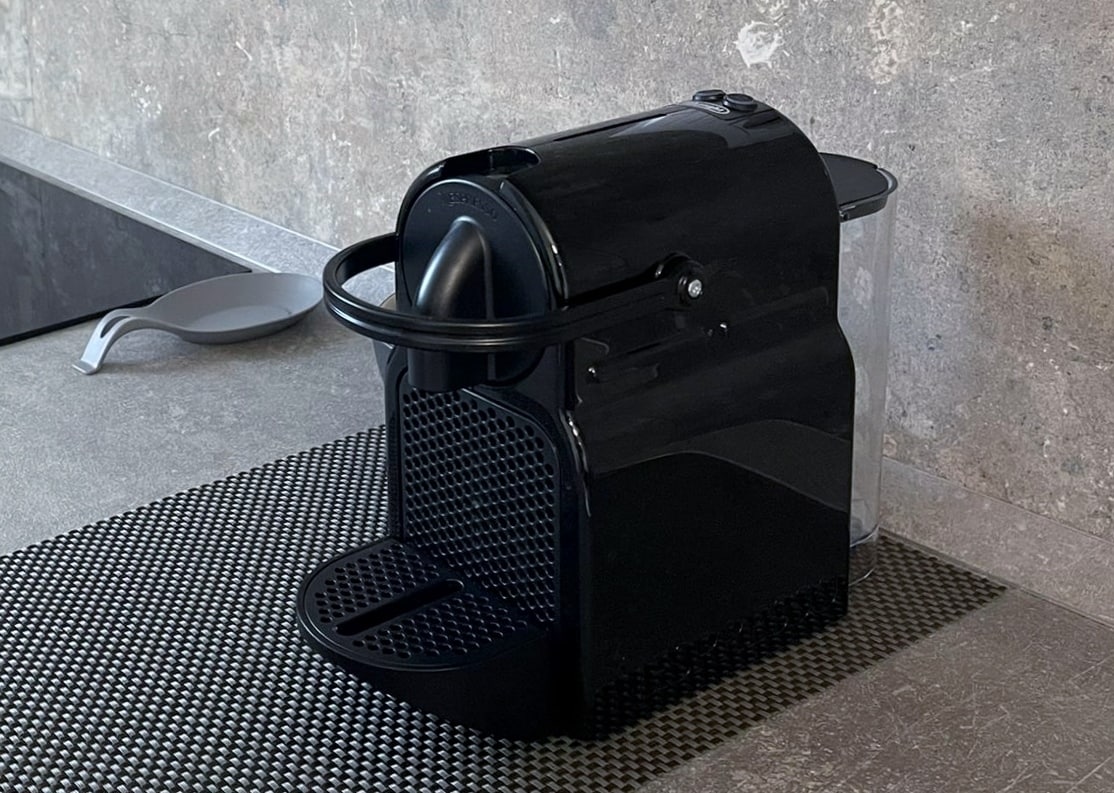 A black Nespresso coffee machine is positioned on a textured mat. The machine features a streamlined design with a prominent water reservoir, positioned against a minimalist grey countertop.