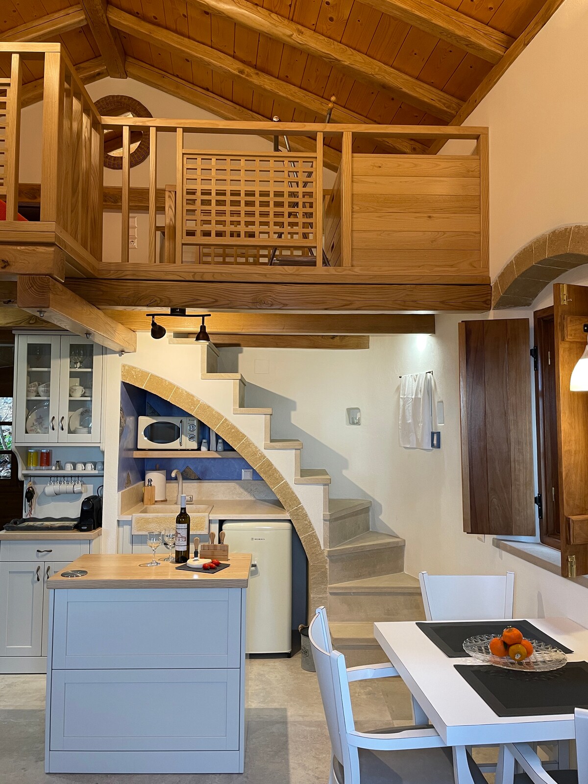 The open-plan living area features a wooden loft above, enhancing the spacious feel of the suite. A dining table with white chairs is positioned beside a kitchen area equipped with modern appliances. Curved stone stairs lead to the loft, illuminated by natural light from window openings.