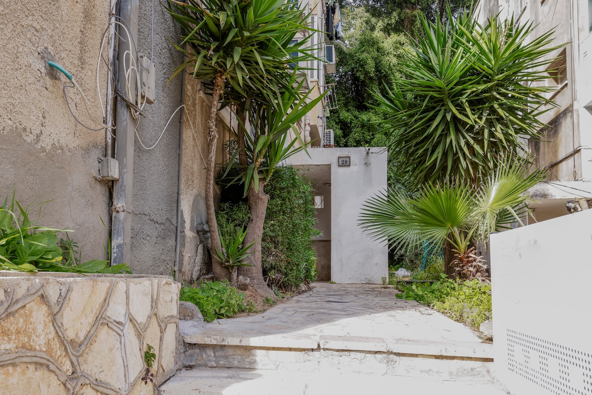 A pathway leads to a bright entrance framed by lush tropical plants and greenery. The walkway features a textured stone surface and is lined with vibrant foliage, providing a welcoming entry to the property.