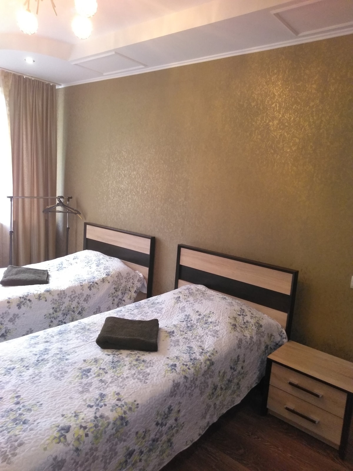 A bright and inviting room features two twin beds with floral-patterned bedding. Each bed is paired with a bedside table. Golden-hued wallpaper adds warmth to the space, while natural light enters through a window covered by a light curtain.
