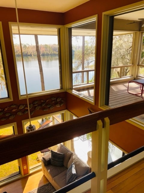 A spacious living area features large windows that provide scenic views of the river and surrounding nature. A comfortable couch is positioned near the windows, inviting relaxation while the sunlight brightens the room. The balcony outside extends the living space, showcasing the peaceful waterfront setting.