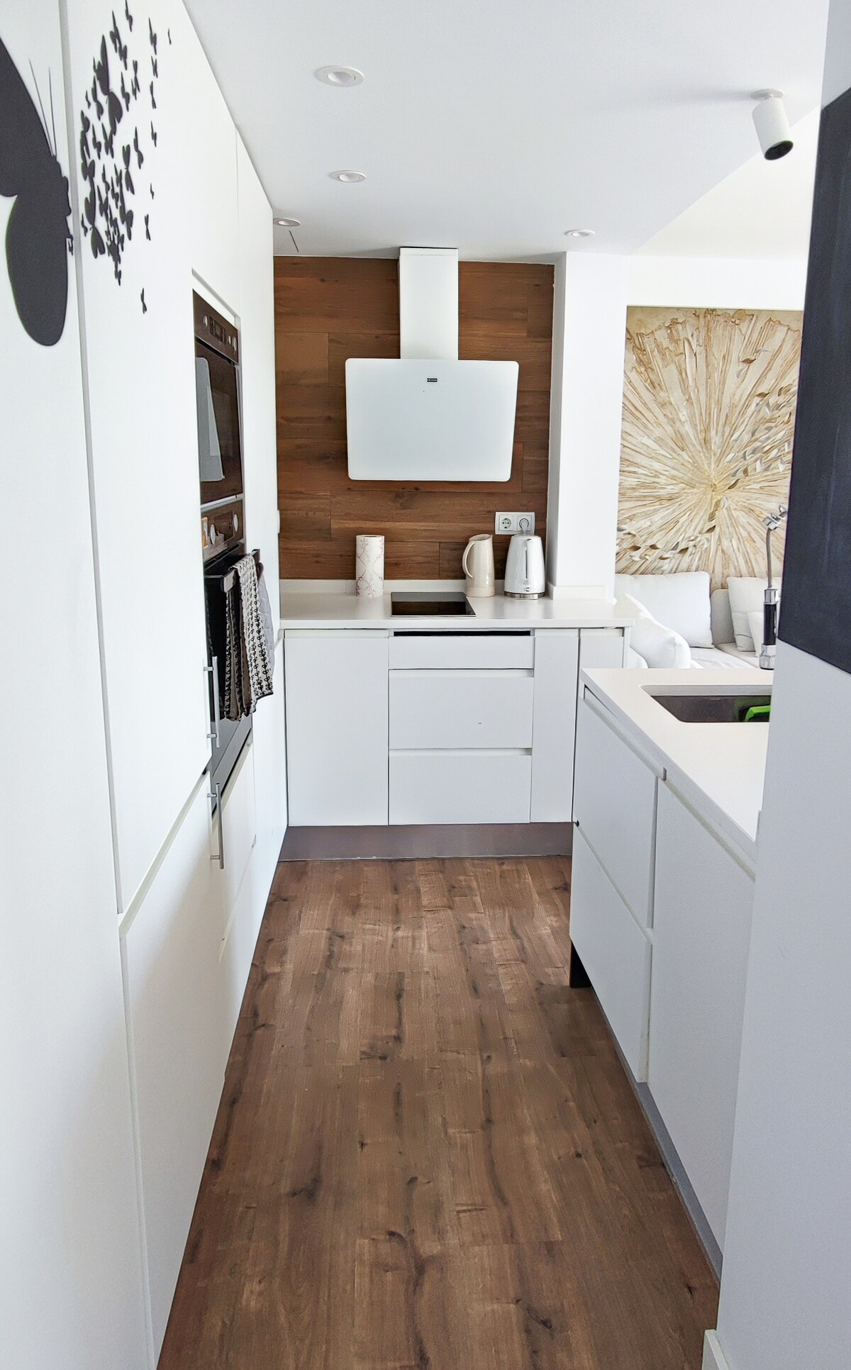 A modern kitchen space features sleek white cabinetry and wooden accents. Stainless steel appliances, including an oven and kettle, are visible. Ample counter space surrounds a sink, and a minimalist design contributes to a clean, uncluttered appearance.