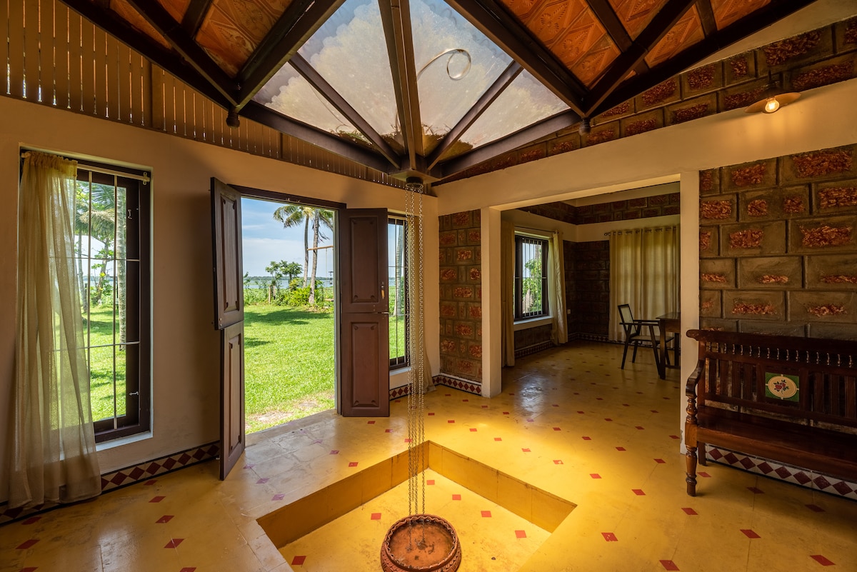 An open living space features large windows providing views of the lush garden and backwaters. The room includes a decorative hanging feature above a circular area on the yellow floor. Light-colored walls and wooden accents enhance the serene atmosphere.
