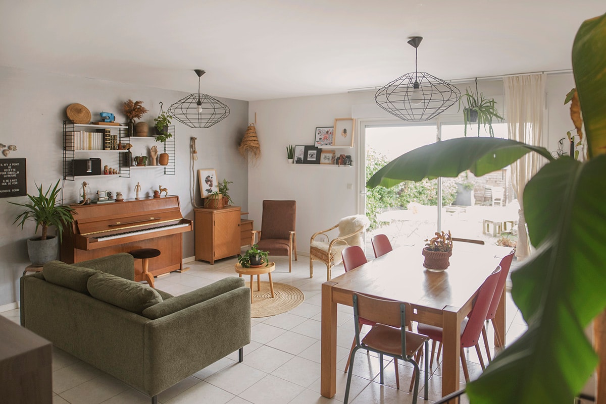 A bright living area features a mix of greenery and warm furnishings. A piano sits beside a wooden cabinet, while a dining table with red chairs is positioned near expansive windows. Light filters through sheer curtains, illuminating the space and connecting it to the outdoors.