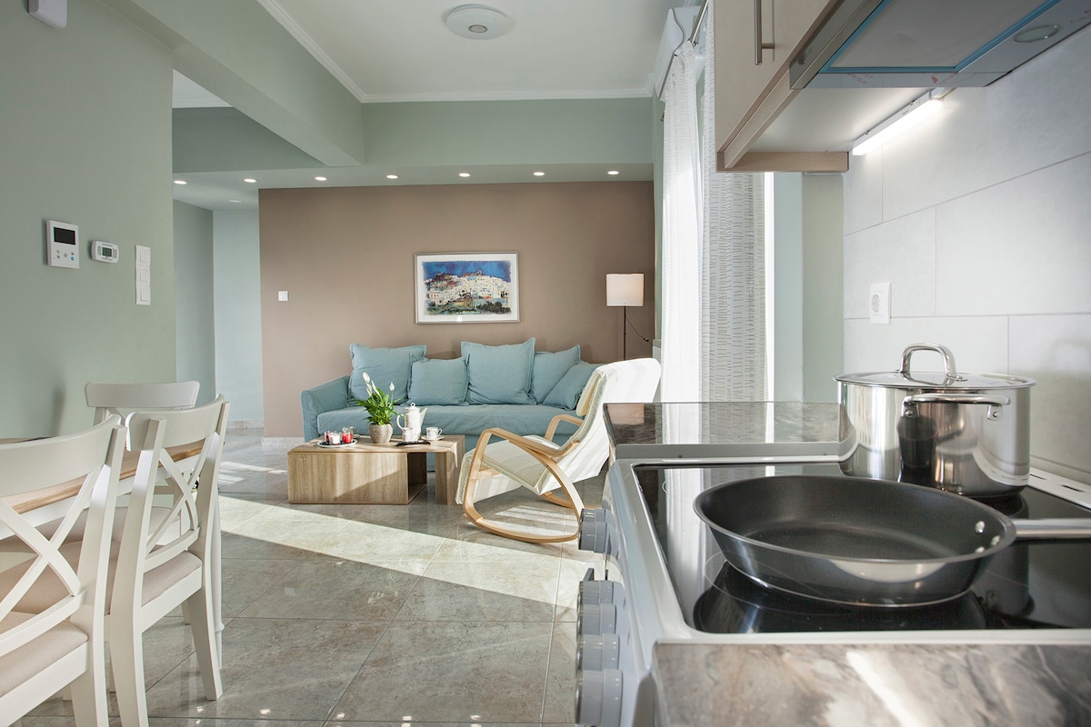 A modern living area features a comfortable seating arrangement with a light blue couch and a stylish wooden coffee table. A spacious kitchen is visible, with cookware neatly arranged on the stove. Natural light illuminates the room, enhancing the serene atmosphere.