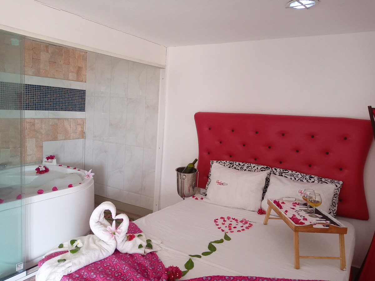 A cozy hotel room features a spacious bed with decorative pillows. A red tufted headboard provides a bold contrast. A luxurious jacuzzi is visible through glass partitions, while a bedside tray holds drinks. Decorative flower petals adorn the bed and floor, enhancing the room's ambiance.