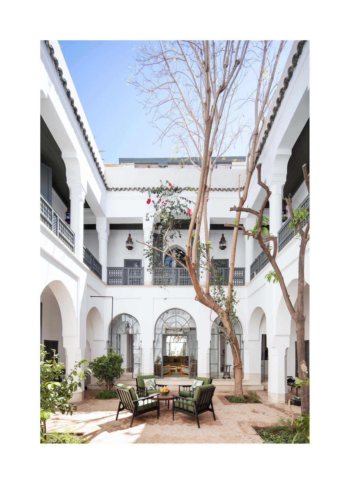 A tranquil patio area is framed by whitewashed walls and arches, showcasing lush greenery and a central tree. Comfortable seating is arranged below the tree, inviting relaxation. Natural light floods the space, enhancing the serene atmosphere.
