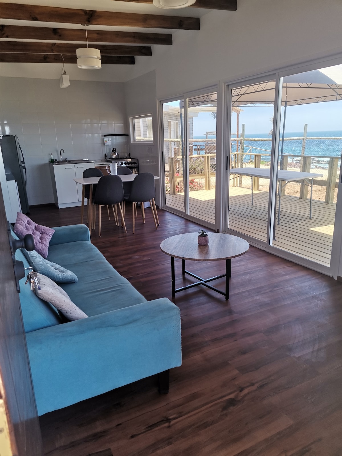 A bright, open living space features a comfortable blue sofa and a small round table. The kitchenette is visible in the background, with modern appliances and dining chairs arranged nearby. Large glass doors lead to a deck with ocean views, allowing for natural light to fill the area.