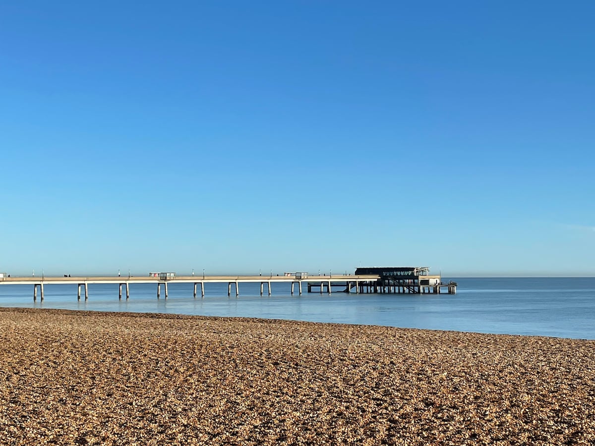 A serene seaside view features a pebbled beach leading to a wooden pier extending over calm waters. A clear blue sky provides a bright backdrop, enhancing the tranquil atmosphere of the coastal setting.