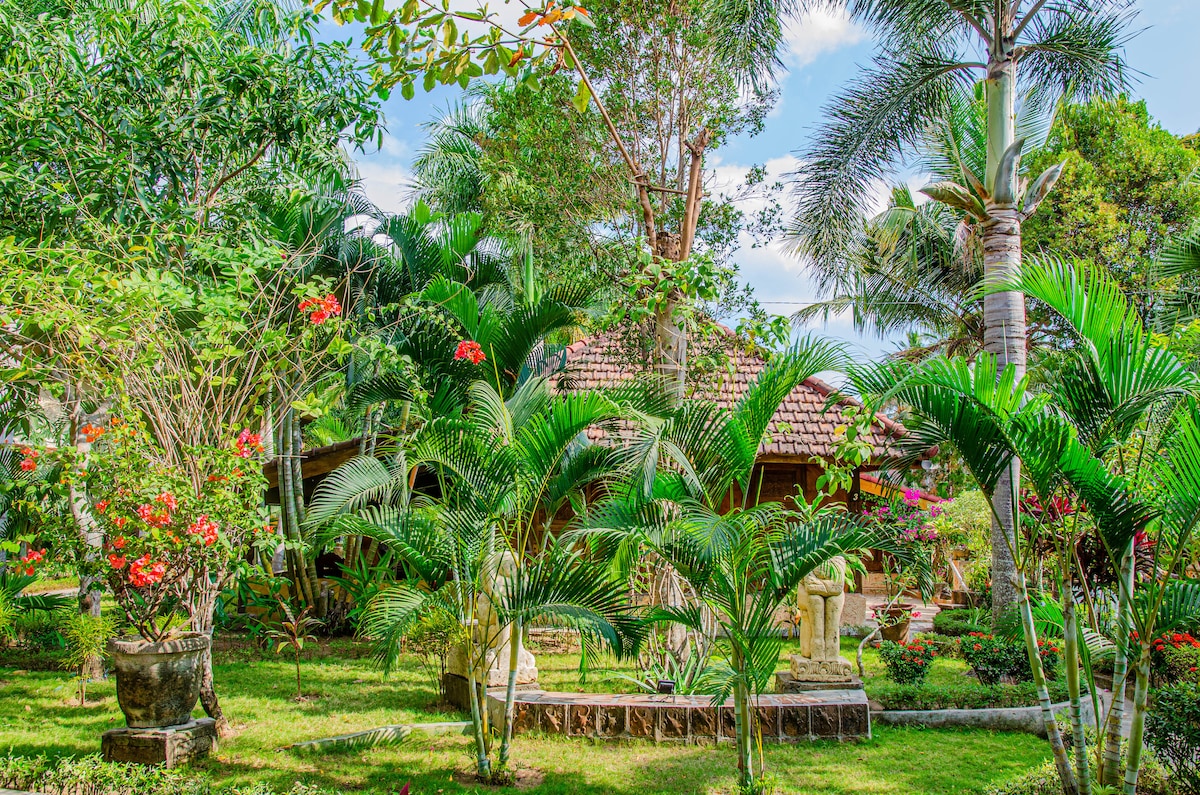 Lush tropical garden is filled with vibrant greenery and colorful flowering plants. Statues and stone decorations can be seen nestled among palm trees and various plants, creating a serene and inviting outdoor space. A charming wooden building is partially visible in the background.