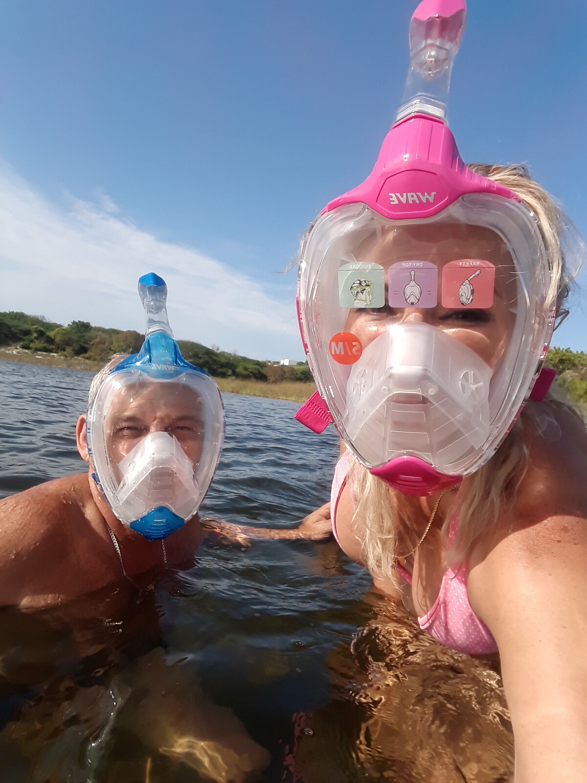 Two individuals are seen enjoying a day in the water, wearing colorful snorkeling masks. The serene, clear water reflects the blue sky above, while greenery is visible in the background, creating a relaxed and engaging outdoor experience.